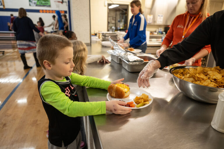 A young Salem Lutheran School student receives a lunch plate with a cheeseburger, chips, and fruit from staff members serving a meal in the school gymnasium.